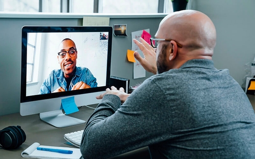 In an organized office cubicle speckled with post-it notes, an office associate sits and makes hand gestures while speaking with a colleague over a video call on his Mac desktop.