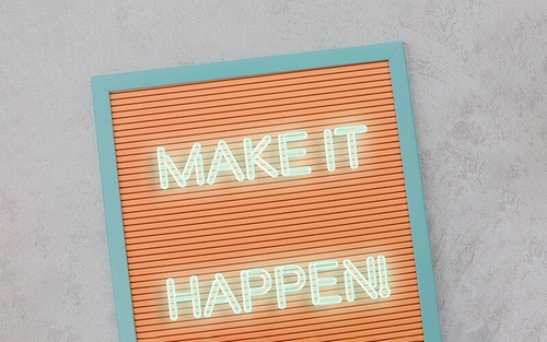 A brown letter board with light blue trim has the saying, 'Make it happen' on it in neon white lettering.