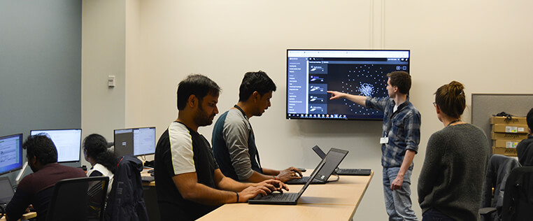 A Schneider engineering associate gestures at data on a large monitor while several colleagues work at laptops and desktops in the room