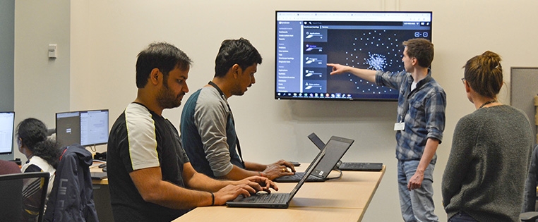 A Schneider engineering associate gestures at data on a large monitor while several colleagues work at laptops and desktops in the room