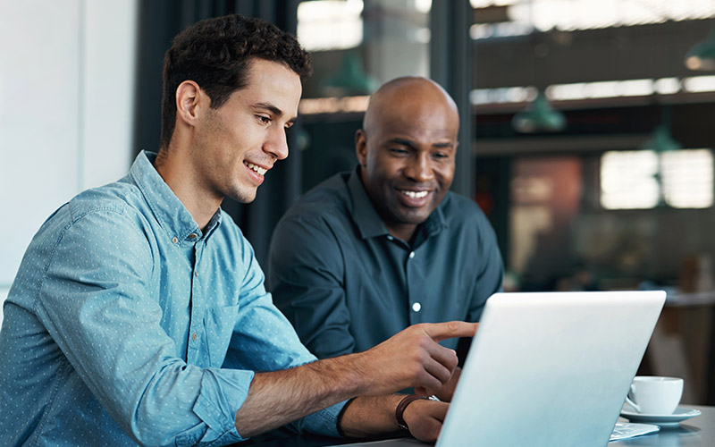 Two men sitting next to each other with a table looking at a computer screen and smiling.