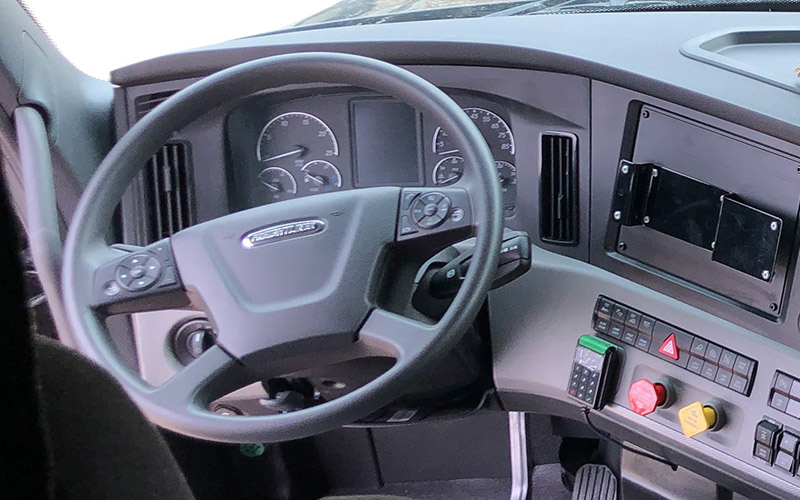 An interior view of a semi-truck, showcasing the grey steering wheel and dashboard.