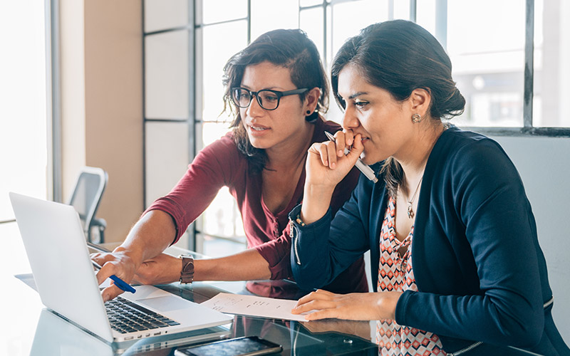 Two women sitting at a desk and looking at a computer while one woman points to the screen.