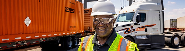 A Schneider Intermodal truck driver at the rail yard standing in front of a Schneider tractor and two Schneider containers