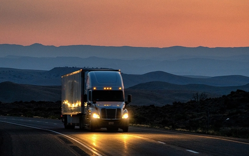 A semi-truck driving on a highway in front of mountains during the sunrise.