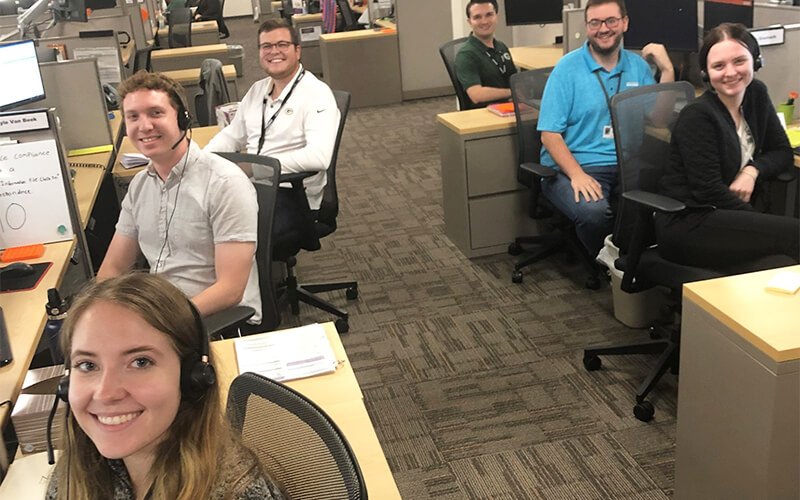 In an office setting are six recruiters who are wearing headsets and sitting at their desks.