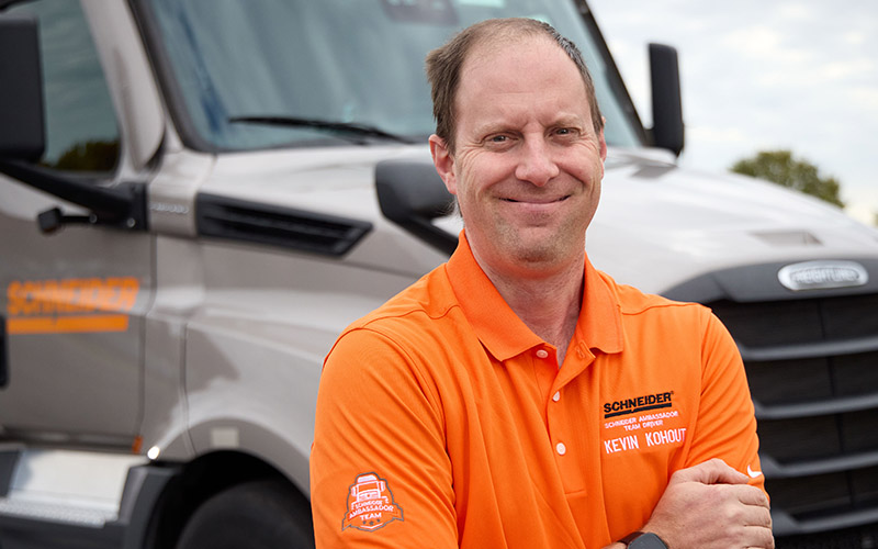 Kevin Kohout standing outside in front of a gray Schneider semi-truck with his arms folded across his chest and smiling straight ahead.