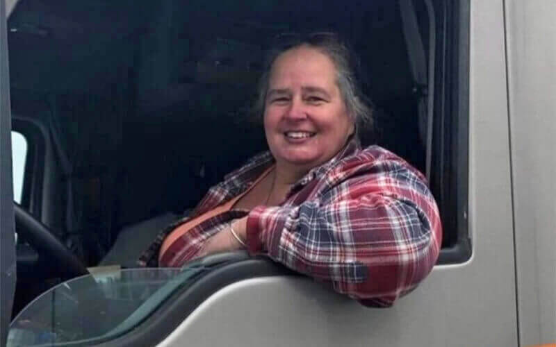 A woman with grey, pulled back hair wears a red and blue plaid shirt and smiles as she leans out of the window.