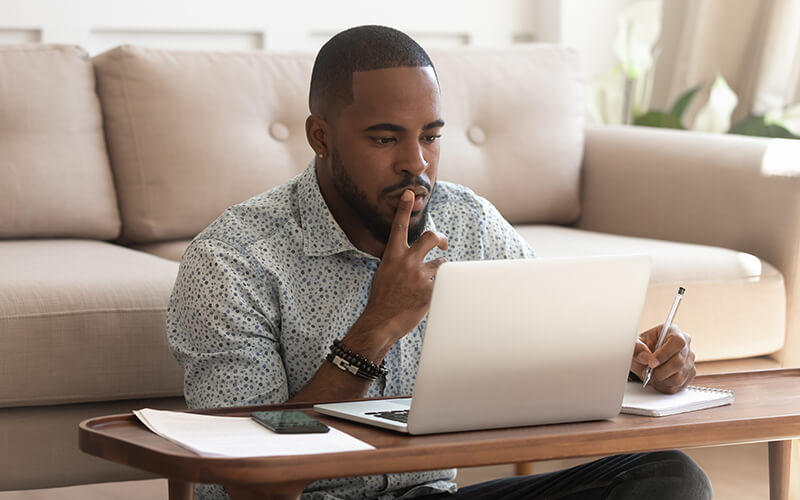 A man is sitting at a desk while working on his laptop. He also has a stack of papers, his phone and a pen and notebook sitting out.