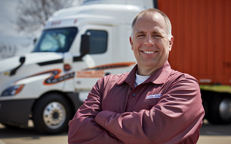 A truck driver wearing a long-sleeve shirt stands outdoors with arms crossed in front of a semi‑truck and a red trailer parked behind.