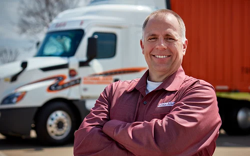 A truck driver wearing a long-sleeve shirt stands outdoors with arms crossed in front of a semi‑truck and a red trailer parked behind.