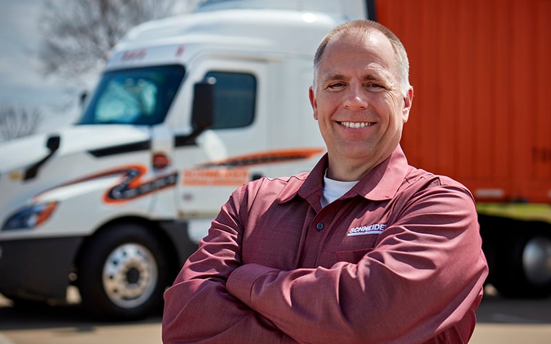 A truck driver wearing a long-sleeve shirt stands outdoors with arms crossed in front of a semi‑truck and a red trailer parked behind.