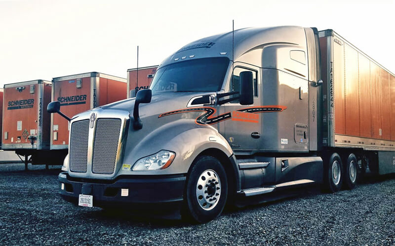 A grey Schneider Kenworth truck with an orange Schneider trailer is parked within a row of other orange Schneider trailers.