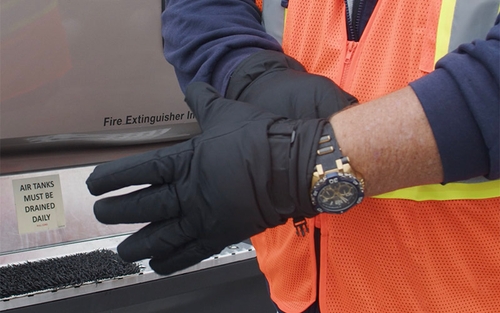 A Schneider truck driver putting on a pair of black canvas work gloves outside of their tractor-trailer.