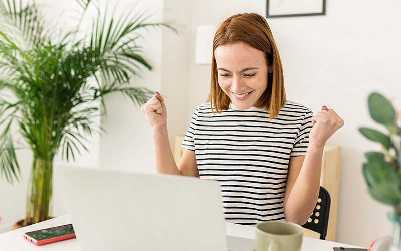 A woman sitting at a table with a laptop in front of her, smiling down at the computer screen with her hands raised triumphantly in the air.