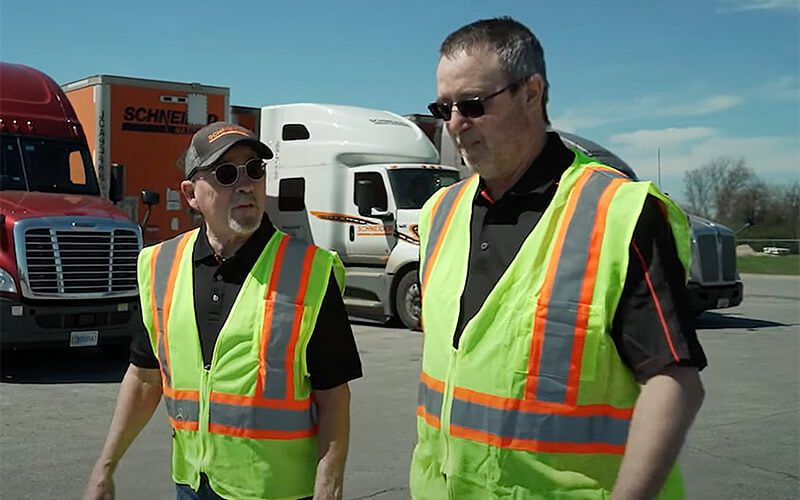 Two Schneider drivers walk side-by-side past semi-trucks parked in a Schneider lot. They are both wearing black shirts, safety vests and sunglasses. The shorter driver on the left is wearing a Schneider hat.