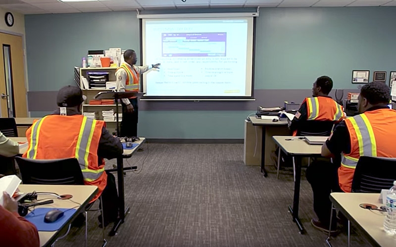 A Schneider Training Instructor wearing a tan shirt and black pants stands in front of a classroom full of truck driving students who are seated and wearing neon safety vests.