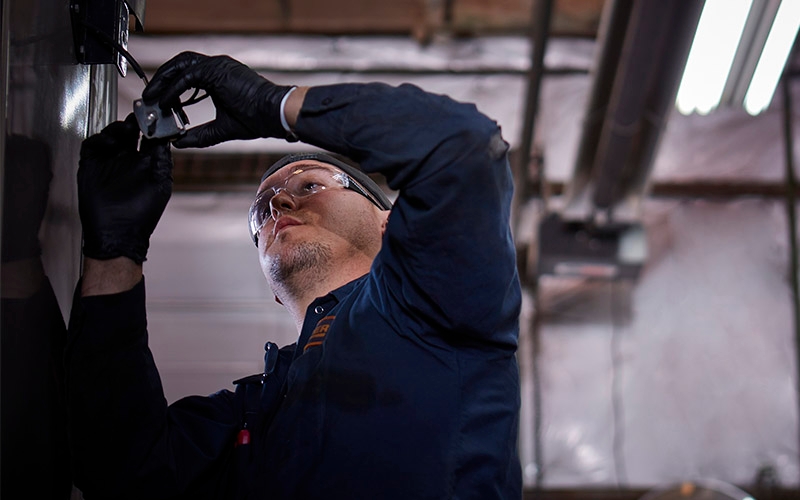 A diesel technician working on a piece of equipment. He's wearing safety glasses, gloves and a long-sleeve uniform.