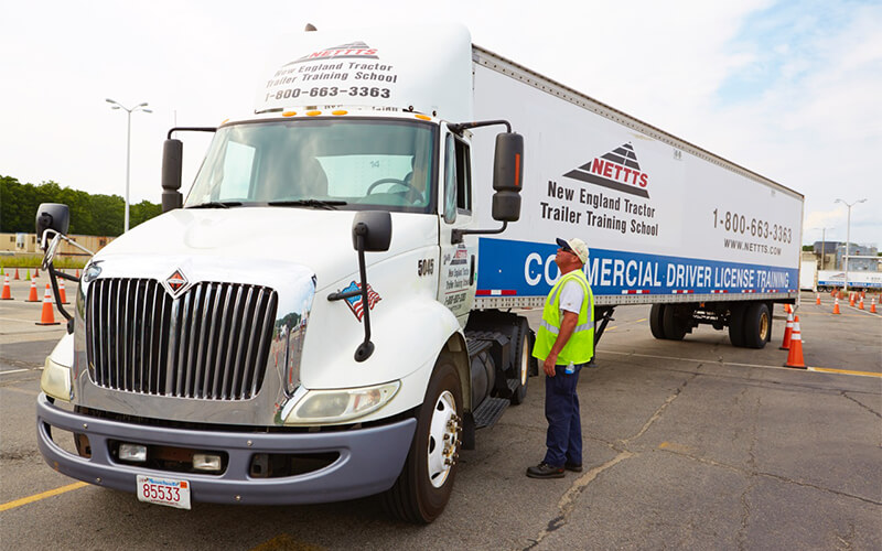 A man in a yellow safety vest and jeans stands in a parking lot and talks to a driver who is backing a white tractor-trailer.