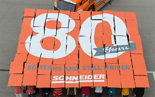 80 Schneider associates stand holding signs over their heads that together create a graphic that reads "80 years and still driven"
