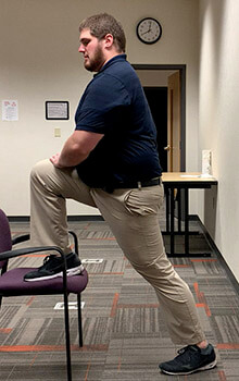 A physical therapist demonstrates stretching his quad by propping his right foot up onto a chair, bending his knee and leaning forward.