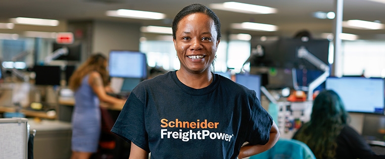 A Schneider associate standing wearing a Schneider FreightPower shirt with several associates working at their desks behind her