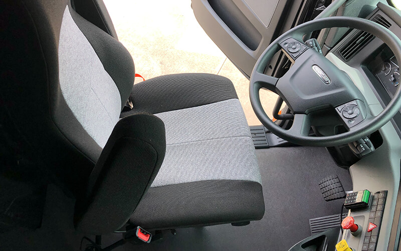 The driver seat of a semi-truck sits empty with the driver-side door open. The seat is grey with black trim.