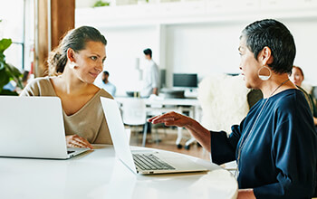 Two women sit at a white, round table with laptops in front of them in an office setting and have a conversation with one another.