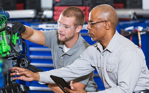 A diesel technician instructor helping a student analyze semi-truck systems. The student is wearing safety eyeglasses and black rubber gloves.