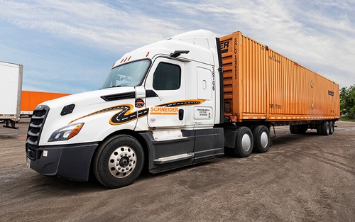 A white Schneider tractor with MirrorEye camera systems, carrying an orange intermodal container is parked at an angle in a dirt parking lot.