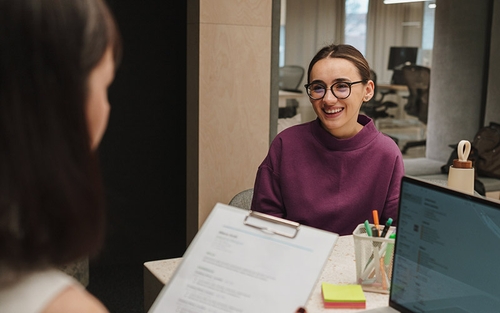A woman sitting at a desk across from a woman with a clipboard, wearing round glasses and a red sweater.