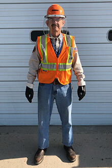A man wears a long sleeves, jeans, gloves, a safety vest and hard hat with a visor.