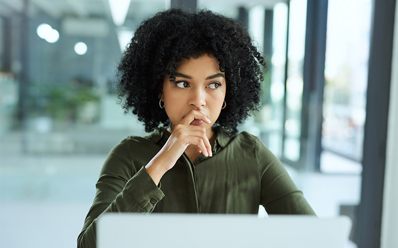 A woman sitting at a table with a laptop in front of her. She's looking to the side and her hand is touching her mouth.