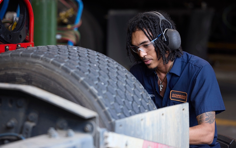 A male Schneider diesel technician squatting down and working on the tire of a semi-truck. He's wearing hearing protection and safety eyeglasses.