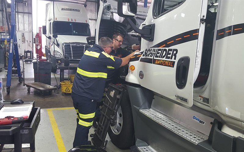 Two diesel technicians work on a while Schneider truck within a shop.