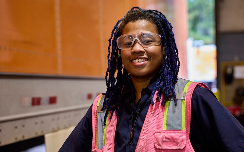 A female diesel technician wearing safety glasses and a safety vest. She's smiling and standing next to an orange Schneider semi-truck.