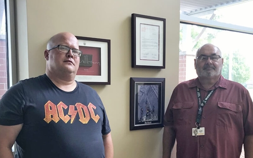 Otis Day and Bob Seidl stand in front of a wall displaying framed 9/11 memorabilia. The frames behind them display a piece of debris from the World Trade Center, a letter of thanks from the NYFD Chief and a front-page news image from 9/11.