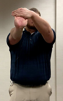 A physical therapist demonstrates stretching his hand by stretching one arm out in front of him with his wrist pointing up and using his other hand to gently bend his fingers back.