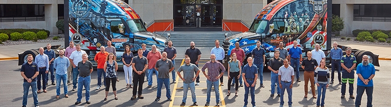 A large group of Schneider office associates that are veterans stand in front of two military wrapped Ride of Pride trucks outside the Schneider corporate office