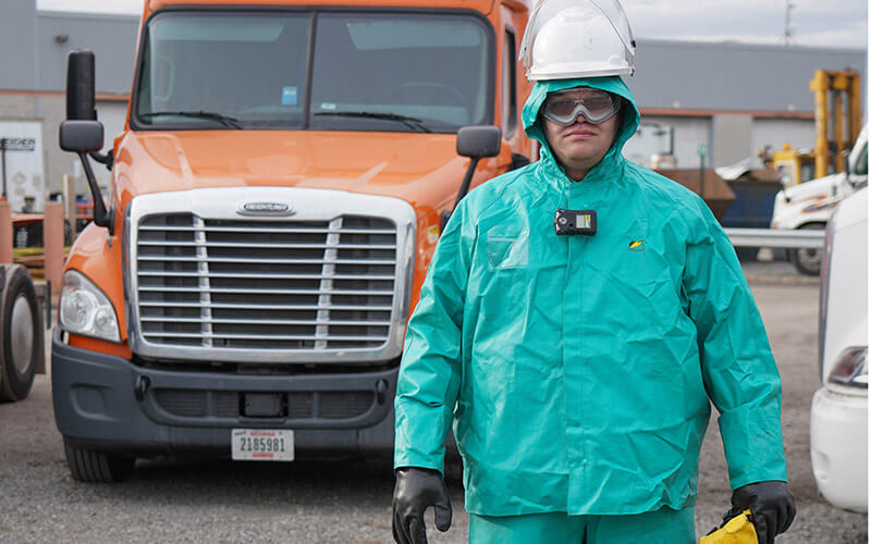 A man wearing a green chemical suit, hard hat, goggles and gloves stands in front of an orange Schneider semi-truck.
