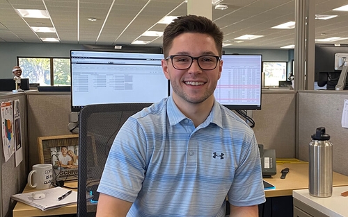 Austin Sylvester, a young man wearing a blue Under Armor polo, sits at his desk that has two desktop computers and a variety of personal items.