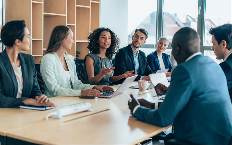 A group of associates are gathered around a table with laptops having a meeting.