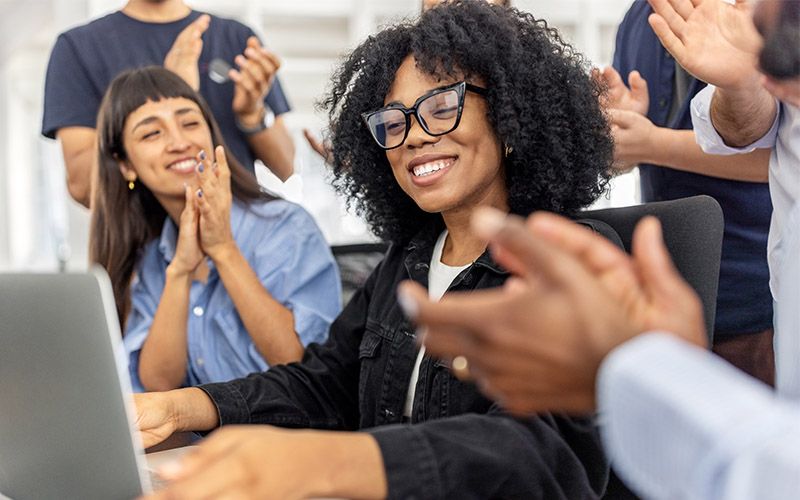 A woman wearing thick black glasses, smiling and sitting around a table while other people applaud her.