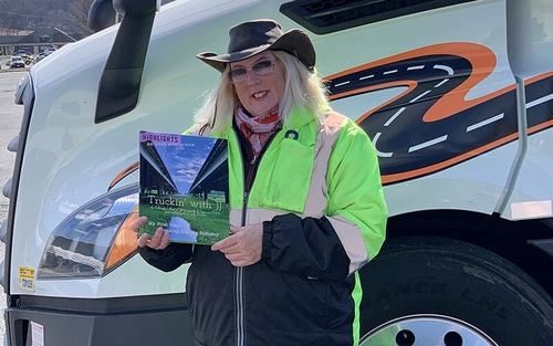 JJ Artsy stands in front of her Schneider truck, wearing a safety jacket and hat, while holding her book, "Truckin' with JJ."