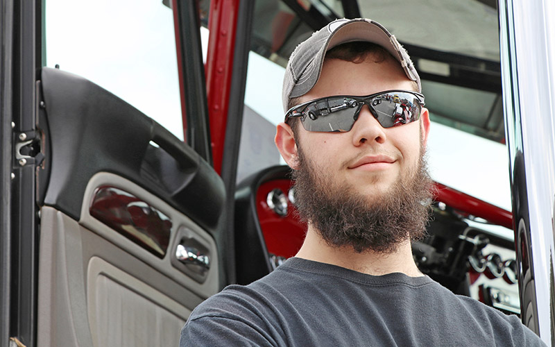 A bearded man wearing sunglasses and a hat stands in front of a semi-truck with it's driver-side door open.