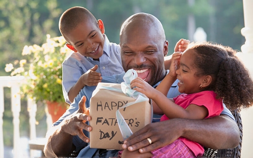 A man smiles and holds his two young children while opening a Father's Day gift.