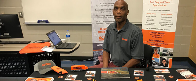 A Schneider field recruiter waits at a career fair booth between two banner displays that outline opportunities for company drivers and owner-operators