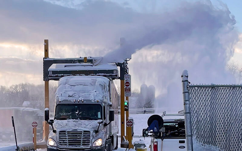 A grey, snow-covered Schneider tractor hauling an orange trailer drives through the YETI snow removal system. The YETI snow remover ejects the snow into the air and away from the truck.