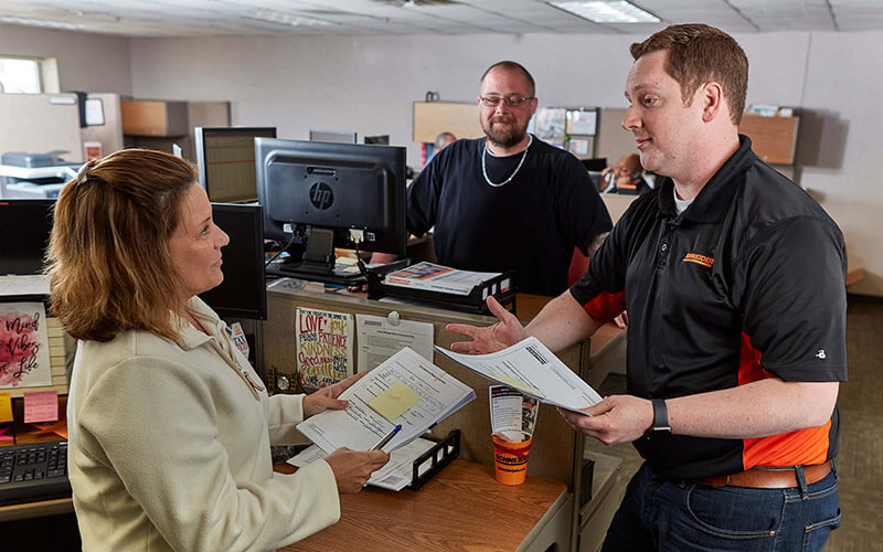 Two men and a woman stand around a desk in an office setting and chat.