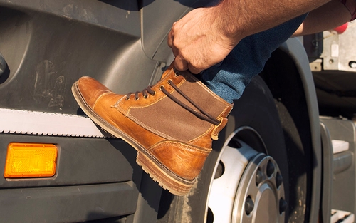 A truck driving lacing up their work boots against the side of the cab of their tractor-trailer.